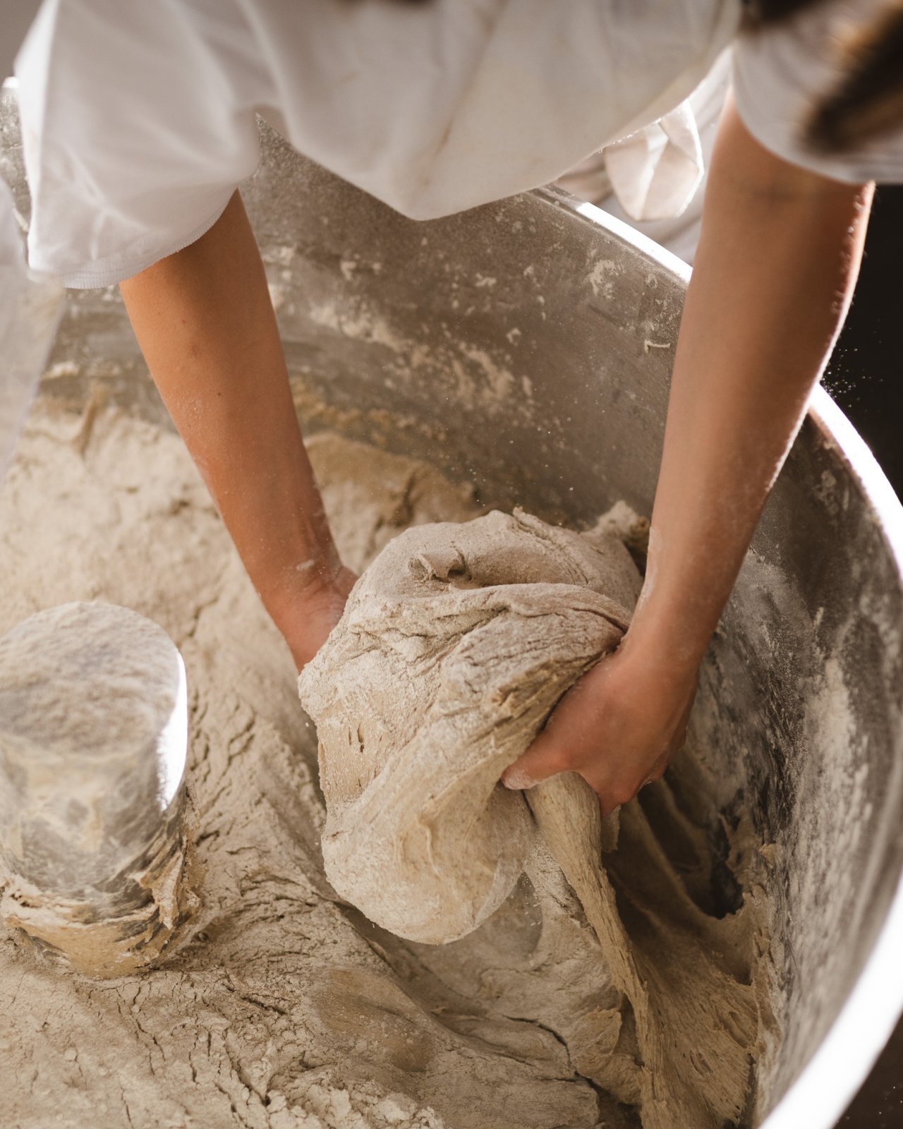 Eine Person knetet Teig in einer gro&szlig;en silbernen Sch&uuml;ssel. Die H&auml;nde sind mit Mehl bedeckt und der Teig hat eine grobe, unregelm&auml;&szlig;ige Form.