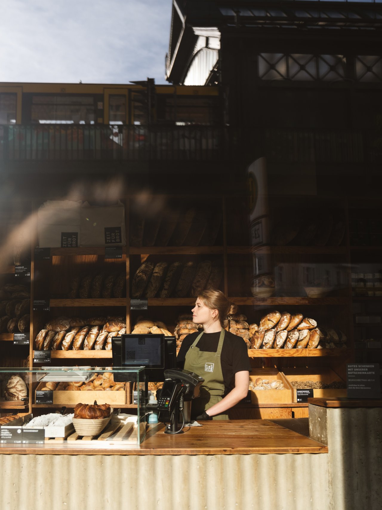 B&auml;ckerei mit Holzregalen voller Brote. Eine Person steht hinter der Theke, tr&auml;gt eine gr&uuml;ne Sch&uuml;rze und hat die H&auml;nde auf der Theke.